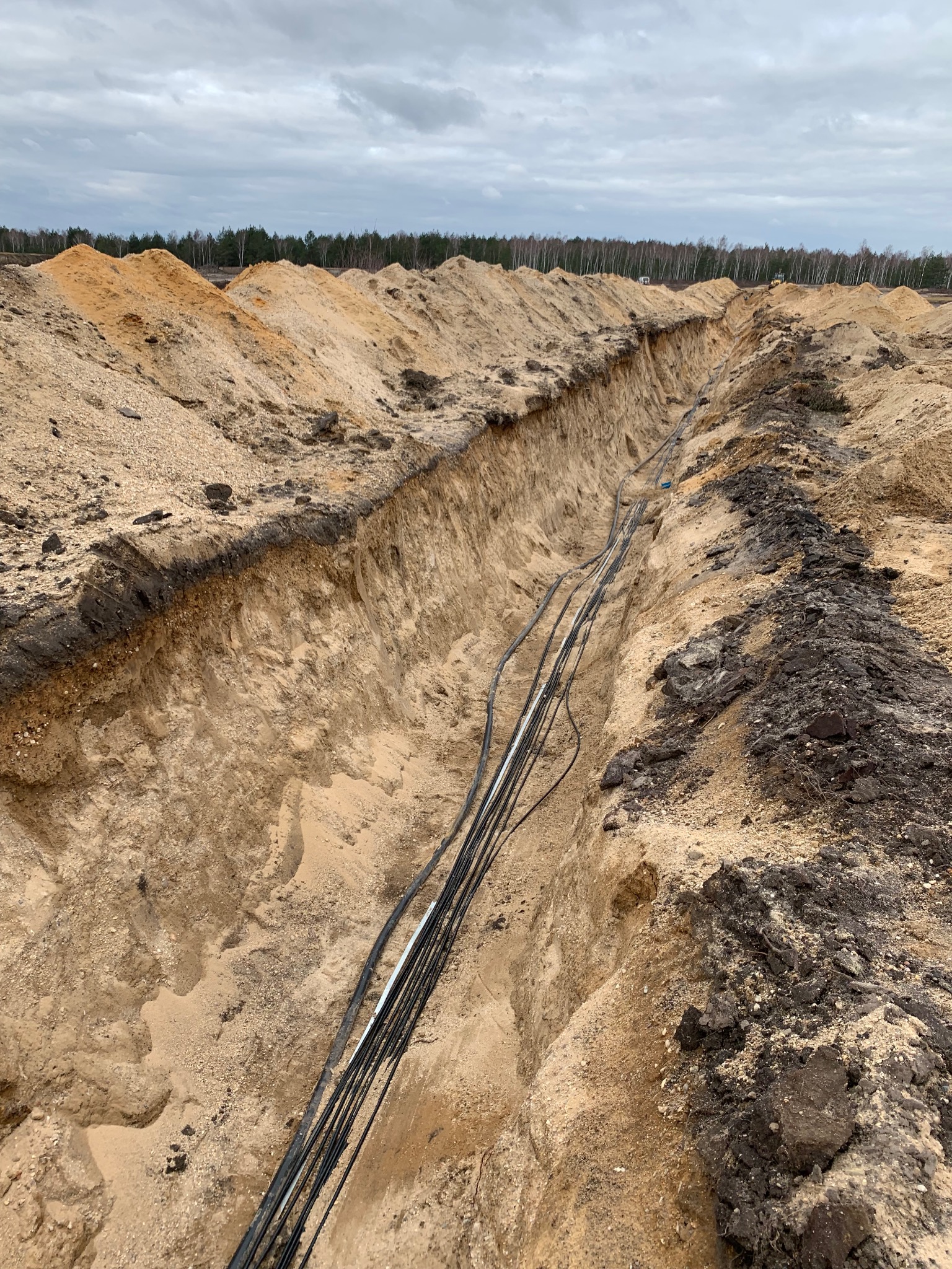 Długi wykop w piaszczystym terenie z ułożonymi przewodami kablowymi, widoczne hałdy piasku i las w tle pod pochmurnym niebem.