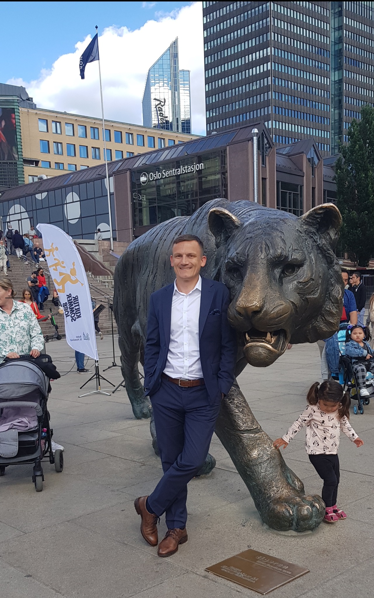 Mężczyzna w garniturze opiera się o pomnik tygrysa na tle Oslo Sentralstasjon, w pobliżu flaga i spacerujące osoby.