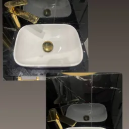 Biała, prostokątna umywalka z pozłacanym korkiem na czarnym blacie z marmuru, widoczne odbicie w lustrze.