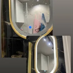 Dwa owalne lustra z podświetleniem LED i złotą ramą, odbijające wnętrze łazienki z czarnymi marmurowymi płytkami, widoczne ślady po myciu na górnym lustrze.