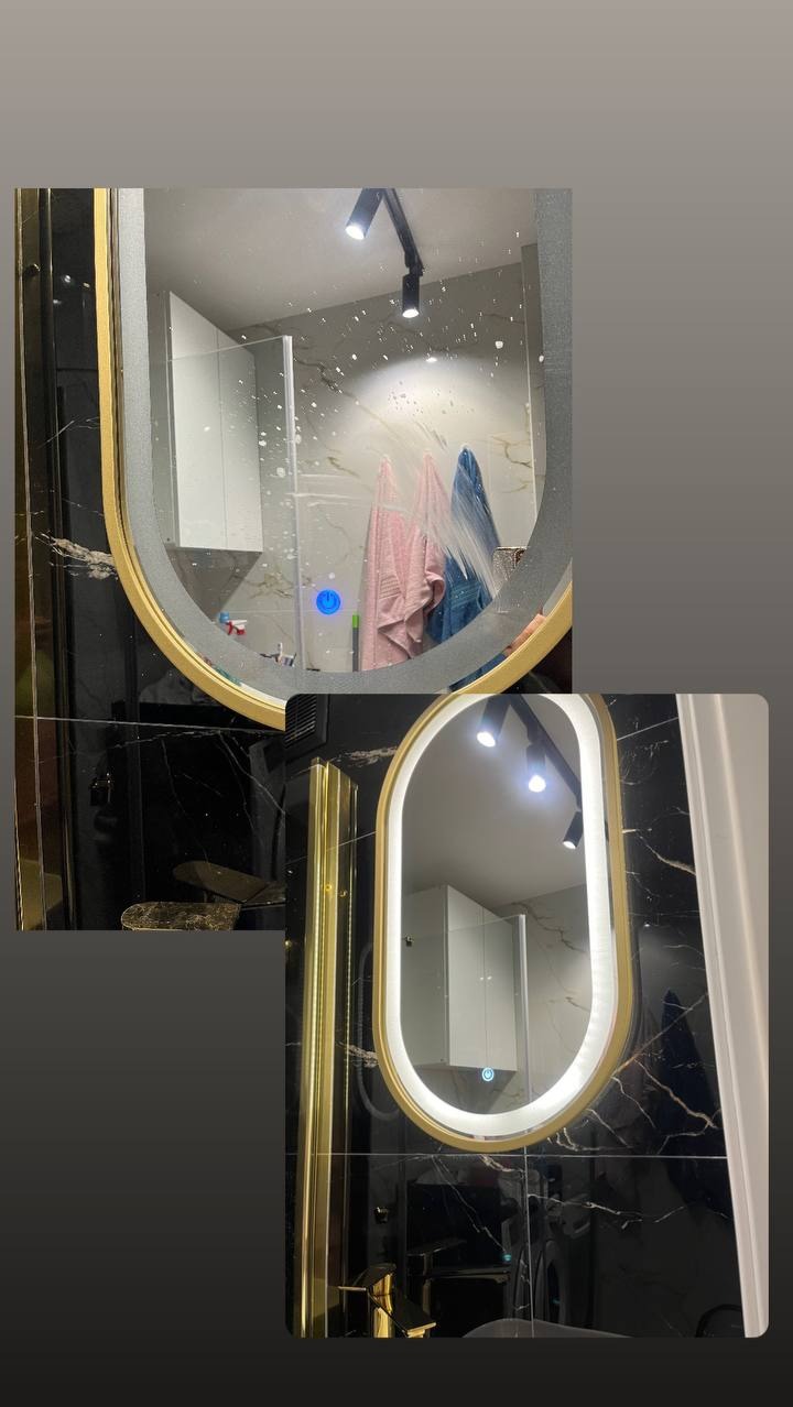 Dwa owalne lustra z podświetleniem LED i złotą ramą, odbijające wnętrze łazienki z czarnymi marmurowymi płytkami, widoczne ślady po myciu na górnym lustrze.