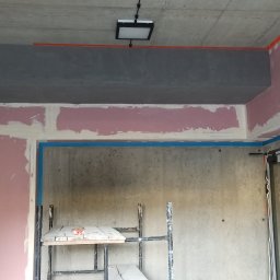 Tomi-BudRem - Surowy pokój w trakcie przygotowania do malowania: ściany z surowego betonu, fragmenty ścian pokryte gładzią, zabezpieczone taśmą malarską, widoczne rusztowanie i skrzynka elektryczna.
