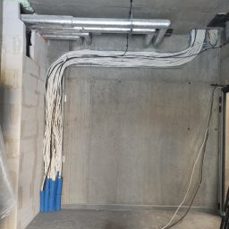 Tomi-BudRem - Instalacja elektryczna w nowym budynku: wiązki kabli zabezpieczone niebieskimi osłonami, prowadzone wzdłuż sufitu i ściany z betonu architektonicznego, widoczne rury wentylacyjne pod stropem.