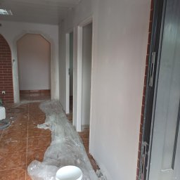 Tomi-BudRem - Wnętrze mieszkania w trakcie remontu: świeżo pomalowane ściany na biało, zabezpieczona folią podłoga z brązowych płytek, widoczny kubeł z farbą marki Śnieżka, przygotowane do malowania futryny...