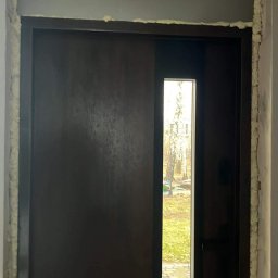 Tomi-BudRem - Montaż brązowych drzwi wejściowych z wąskim przeszkleniem, widoczna piana montażowa w szczelinach ościeżnicy, fragment ściany w szarym kolorze.