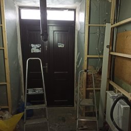 Tomi-BudRem - Wnętrze w trakcie remontu: widoczne drewniane stelaże na ścianach, drabiny, stara stolarka drzwiowa z naklejkami, odsłonięte instalacje, grzejnik oraz elementy wyposażenia budowlanego.