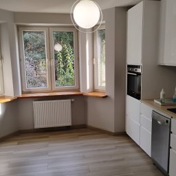Tomi-BudRem - Jasna kuchnia z trzema oknami z drewnianymi parapetami, białymi szafkami, piekarnikiem w zabudowie, zmywarką i drewnianą podłogą.