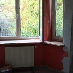 Tomi-BudRem - Pokój w trakcie malowania na kolor terakoty, widoczne dwa okna z zielenią na zewnątrz, grzejnik i wiadro z narzędziami malarskimi.