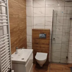 Nowoczesna łazienka z białymi marmurkowymi płytkami, drewnopodobną ścianą z poziomych paneli, wiszącą toaletą i umywalką z szafką, grzejnikiem drabinkowym oraz kabiną prysznicową.