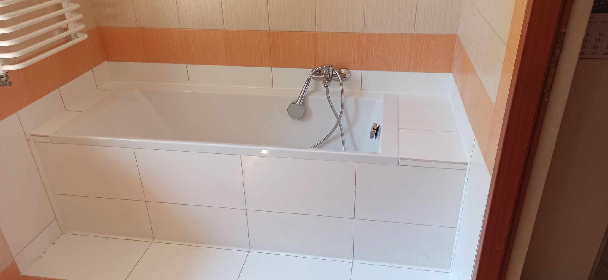 Biała wanna obudowana białymi płytkami, z chromowaną baterią i słuchawką prysznicową, na tle beżowo-pomarańczowych płytek i brązowej framugi drzwi.