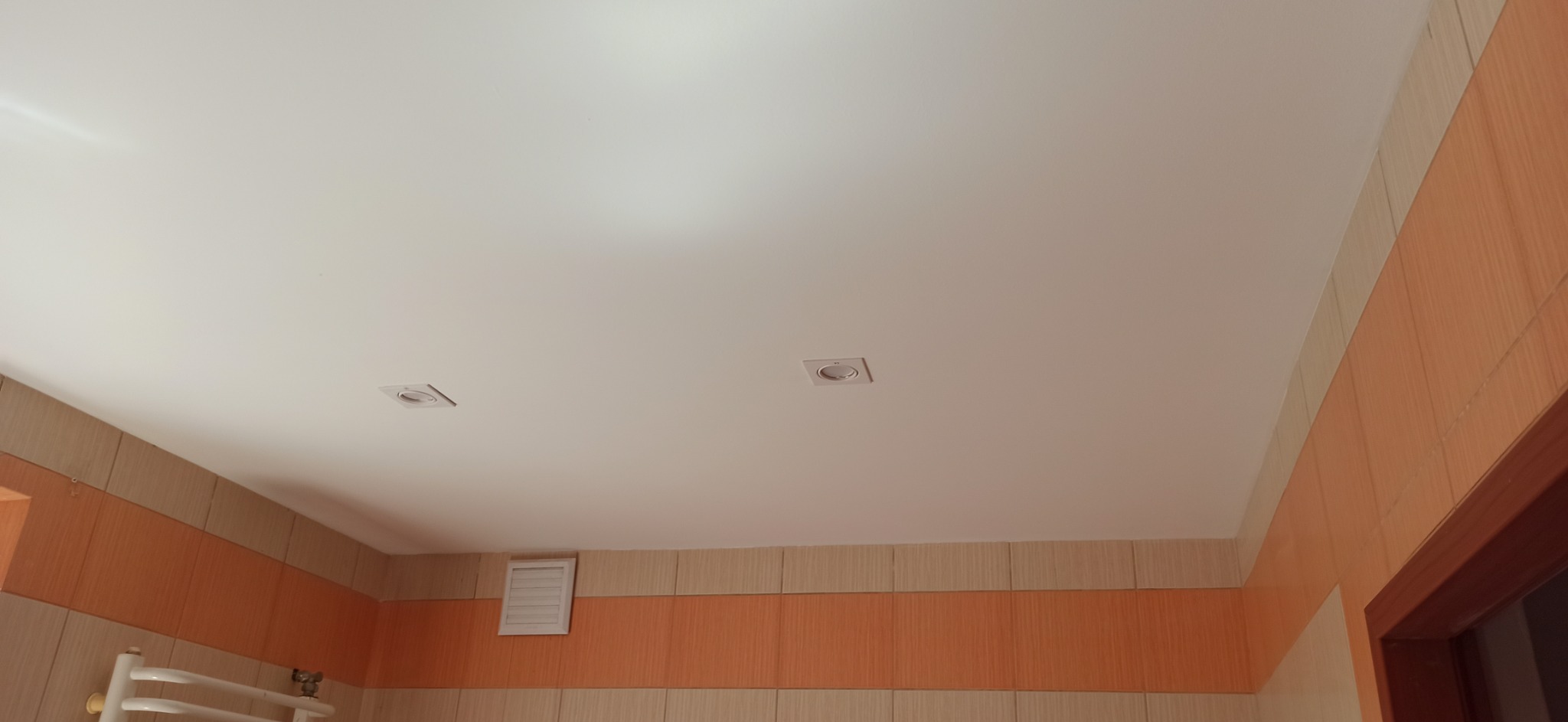 Biały sufit podwieszany z dwoma kwadratowymi lampami halogenowymi w wykończonej łazience z pomarańczowo-beżowymi płytkami i kratką wentylacyjną.