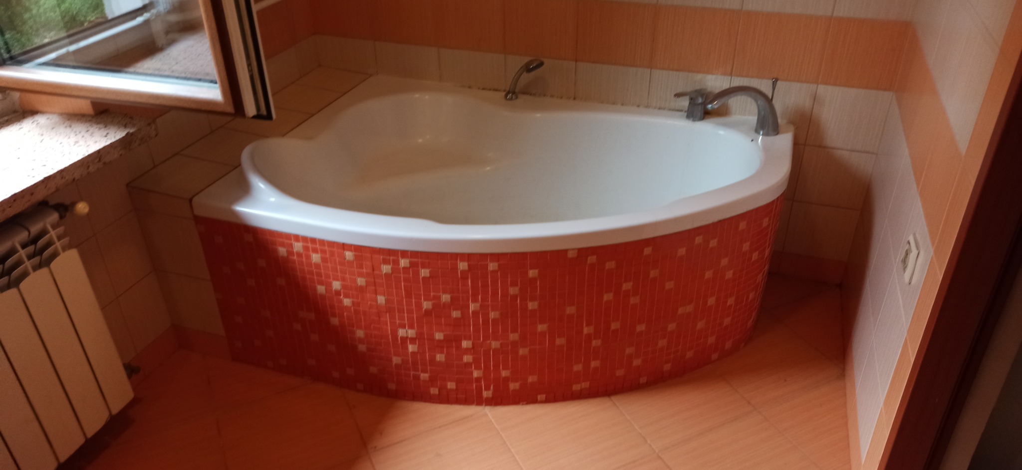 Narożna wanna z hydromasażem obłożona pomarańczową mozaiką w łazience z beżowymi płytkami i grzejnikiem pod oknem.