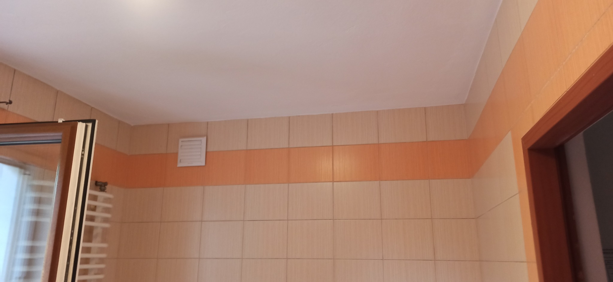 Jasne płytki ceramiczne z pomarańczowym pasem w łazience, widoczny fragment okna i grzejnik drabinkowy.