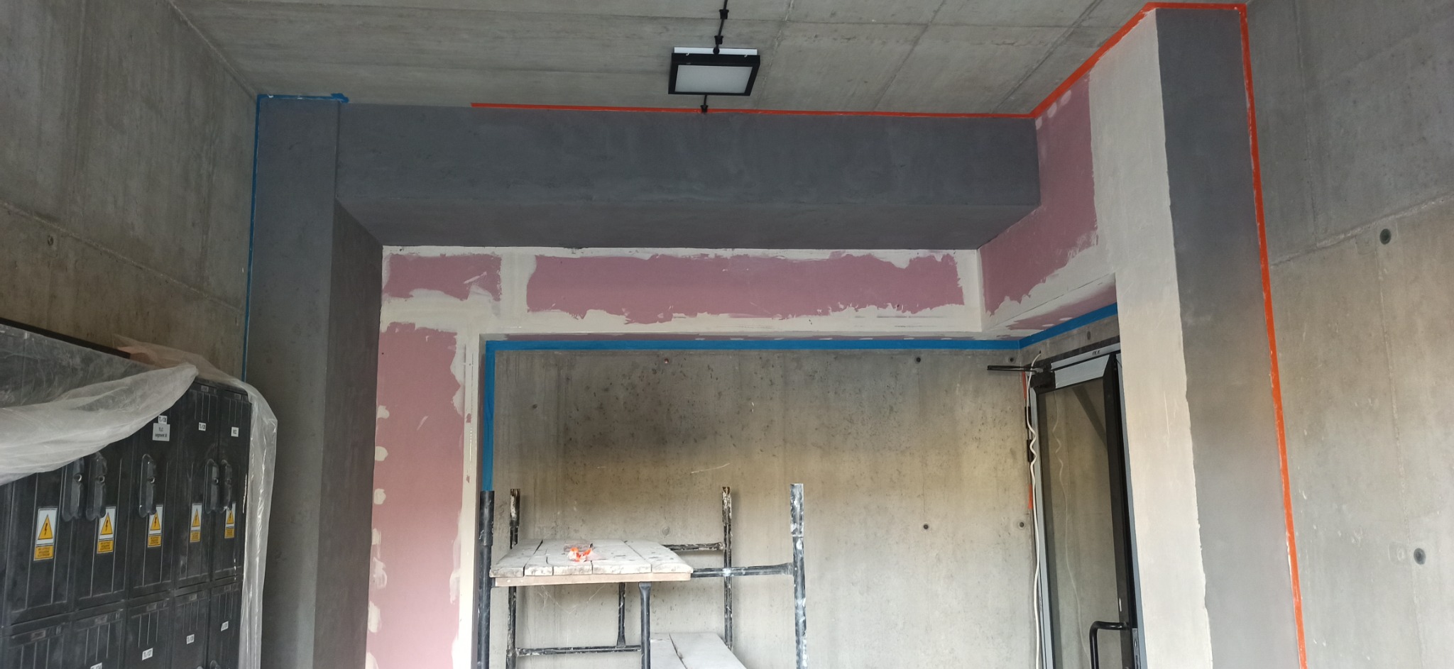 Surowy pokój w trakcie przygotowania do malowania: ściany z surowego betonu, fragmenty ścian pokryte gładzią, zabezpieczone taśmą malarską, widoczne rusztowanie i skrzynka elektryczna.
