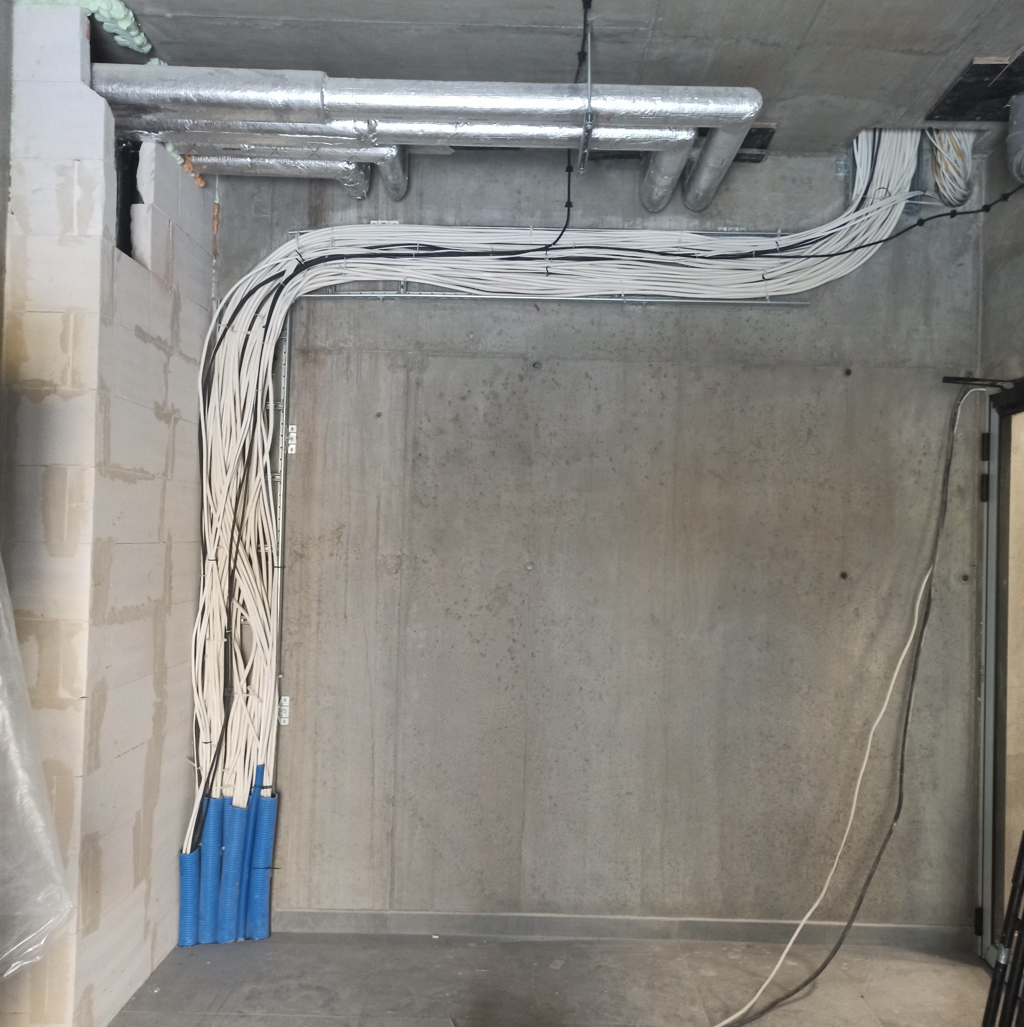 Instalacja elektryczna w nowym budynku: wiązki kabli zabezpieczone niebieskimi osłonami, prowadzone wzdłuż sufitu i ściany z betonu architektonicznego, widoczne rury wentylacyjne pod stropem.