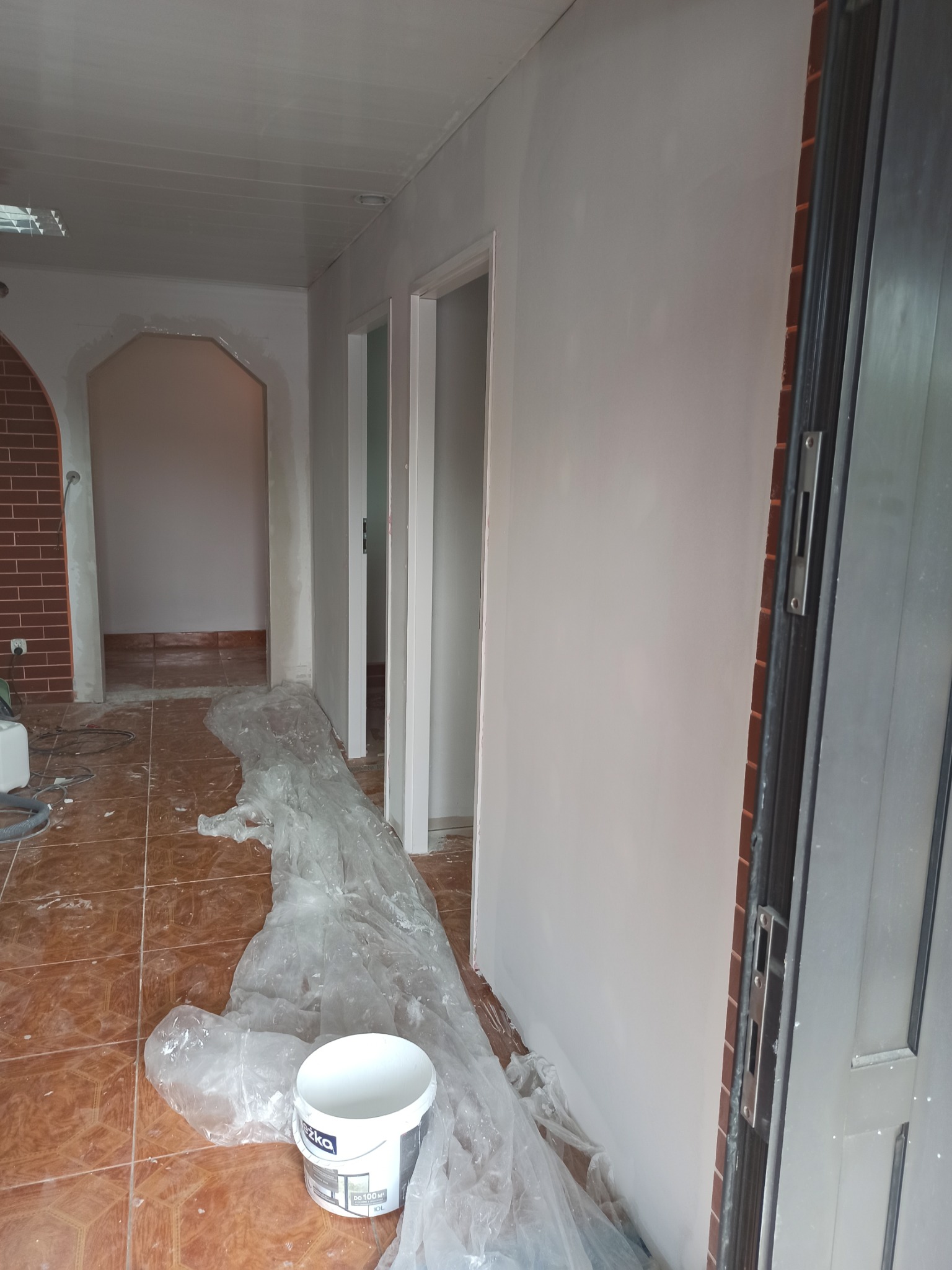 Wnętrze mieszkania w trakcie remontu: świeżo pomalowane ściany na biało, zabezpieczona folią podłoga z brązowych płytek, widoczny kubeł z farbą marki Śnieżka, przygotowane do malowania futryny...