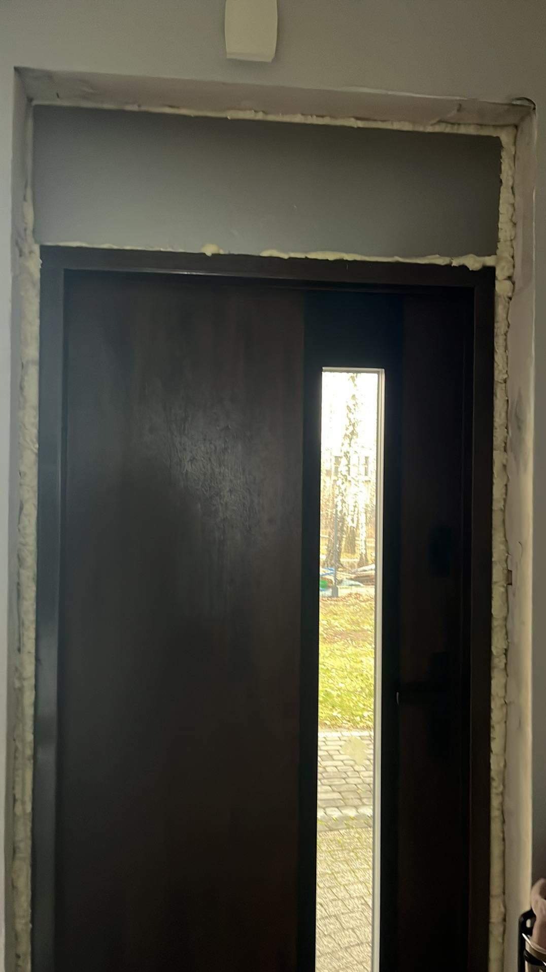 Montaż brązowych drzwi wejściowych z wąskim przeszkleniem, widoczna piana montażowa w szczelinach ościeżnicy, fragment ściany w szarym kolorze.