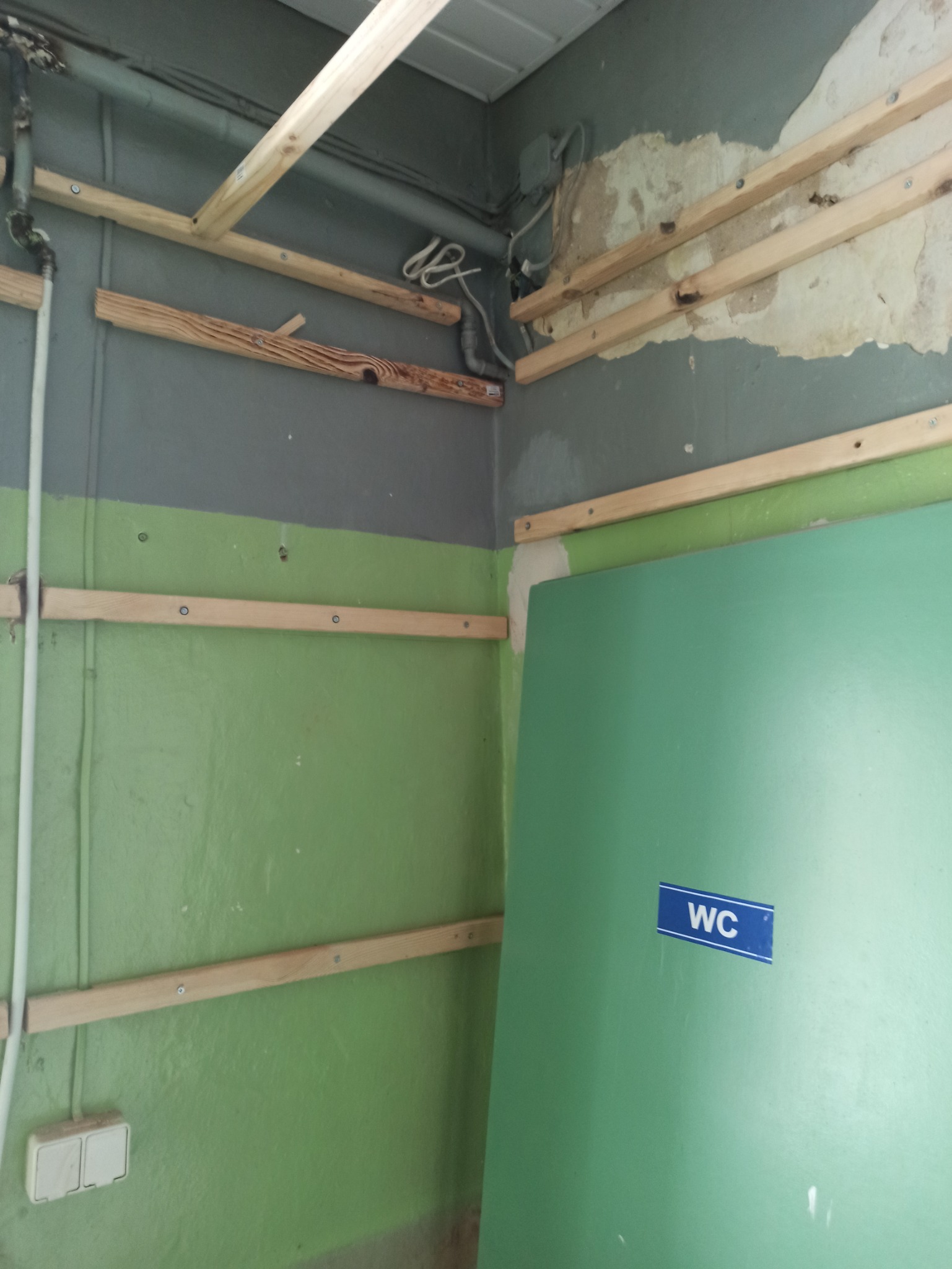 Przygotowanie do remontu łazienki: drewniane listwy mocujące na zielonej i szarej ścianie, widoczne instalacje i drzwi z oznaczeniem WC.