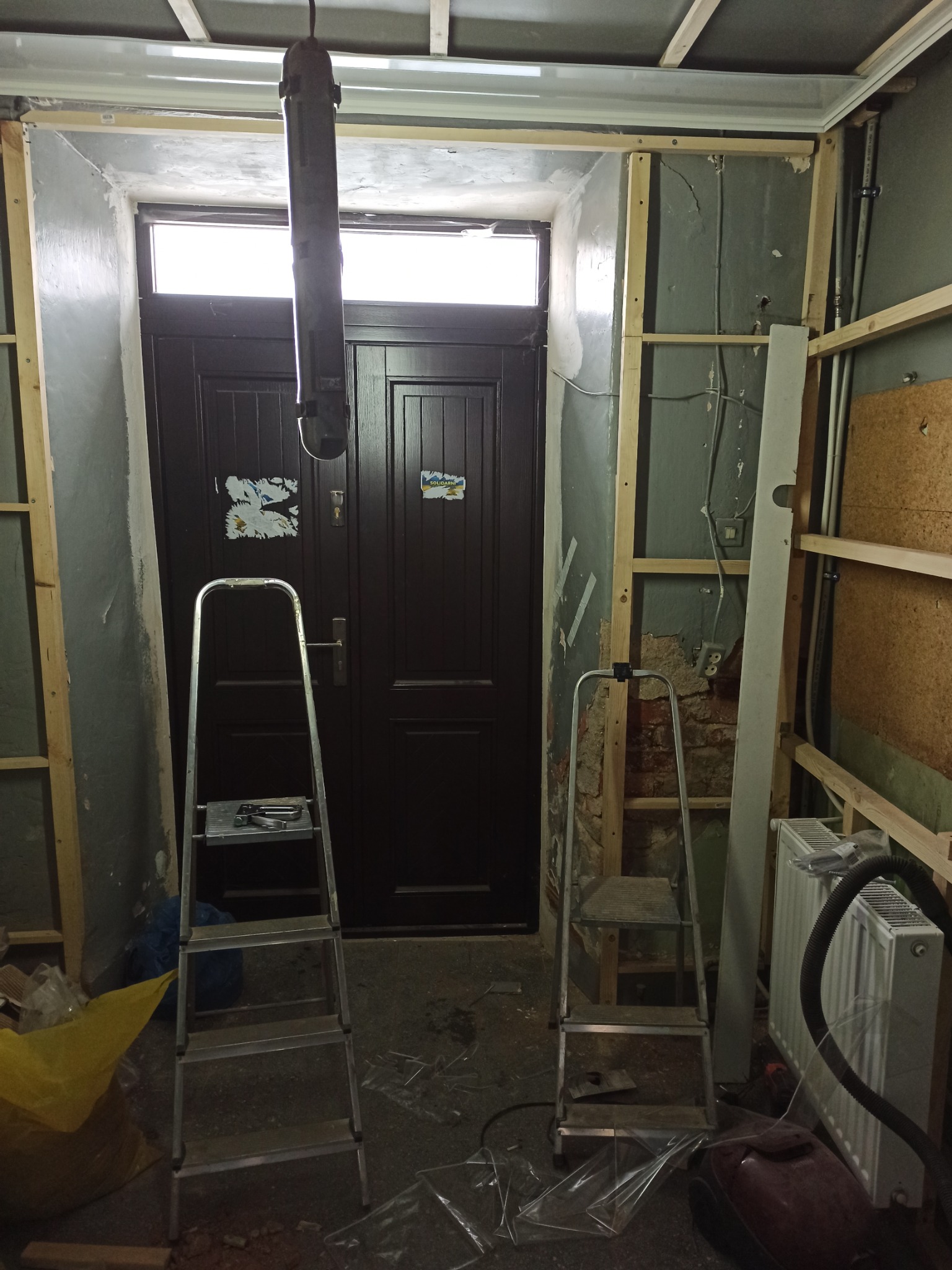 Wnętrze w trakcie remontu: widoczne drewniane stelaże na ścianach, drabiny, stara stolarka drzwiowa z naklejkami, odsłonięte instalacje, grzejnik oraz elementy wyposażenia budowlanego.