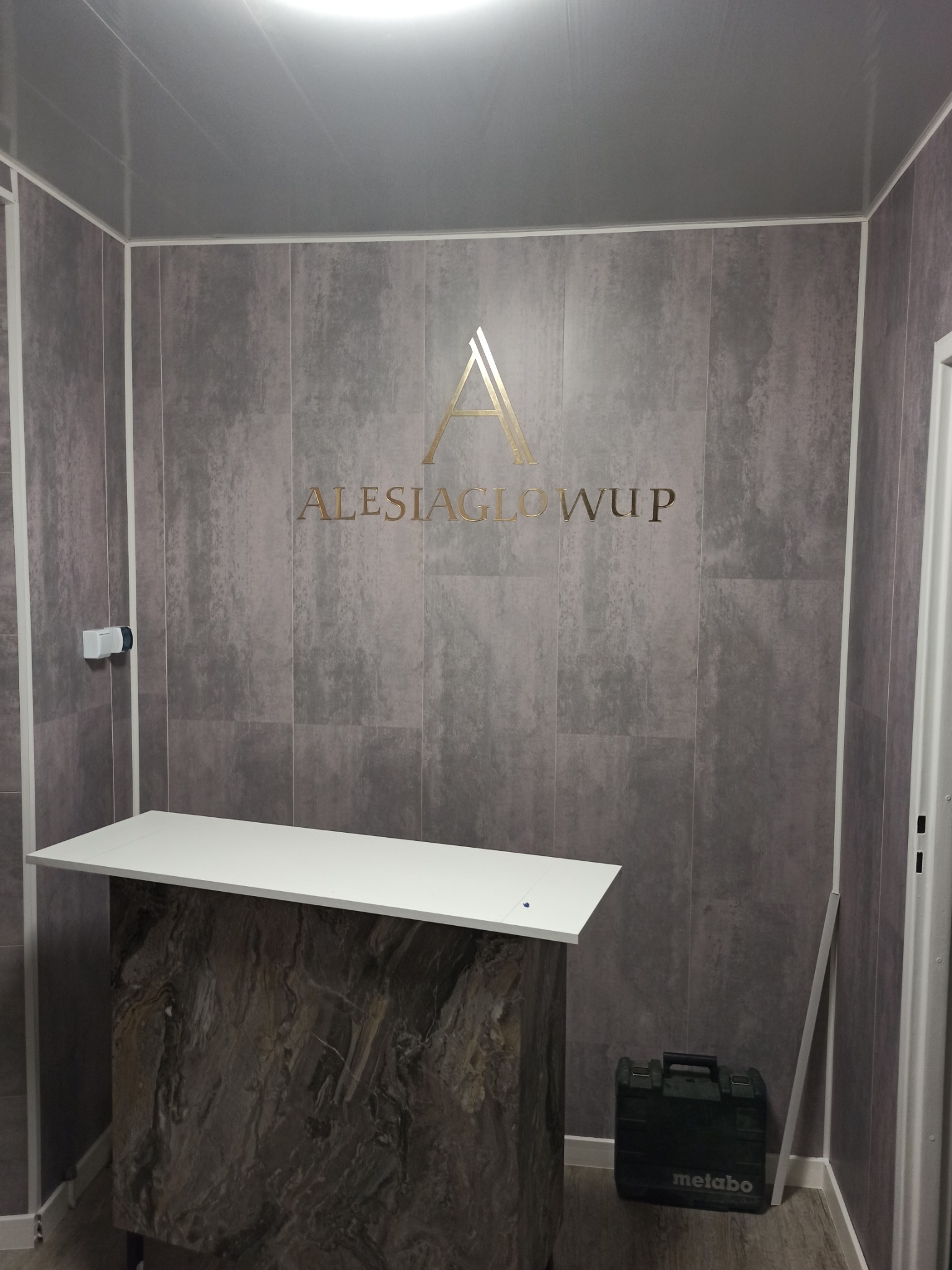 Wykończona łazienka z szarymi płytkami imitującymi beton, białym blatem umywalki na postumencie z płytek imitujących marmur i złotym napisem 'ALESIAGLOWUP' na ścianie.