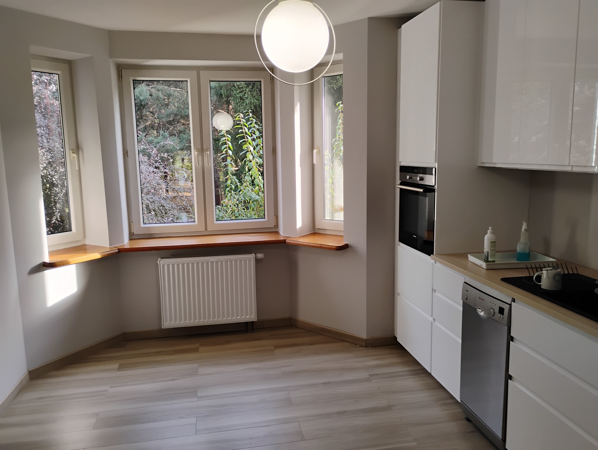 Jasna kuchnia z trzema oknami z drewnianymi parapetami, białymi szafkami, piekarnikiem w zabudowie, zmywarką i drewnianą podłogą.