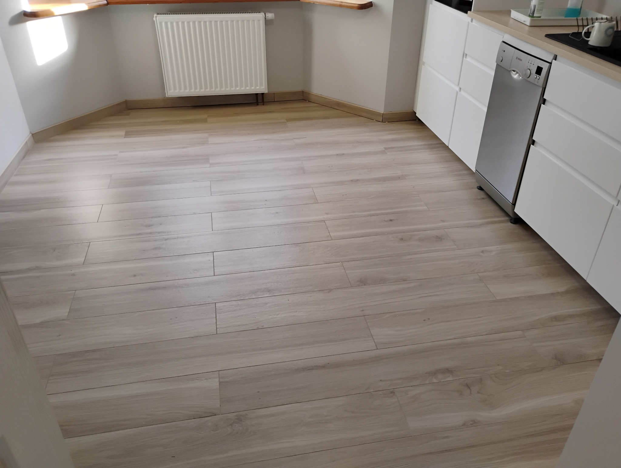 Jasna podłoga z płytek imitujących drewno w kuchni, widoczny fragment białej zabudowy kuchennej ze srebrną zmywarką i grzejnik pod oknem.