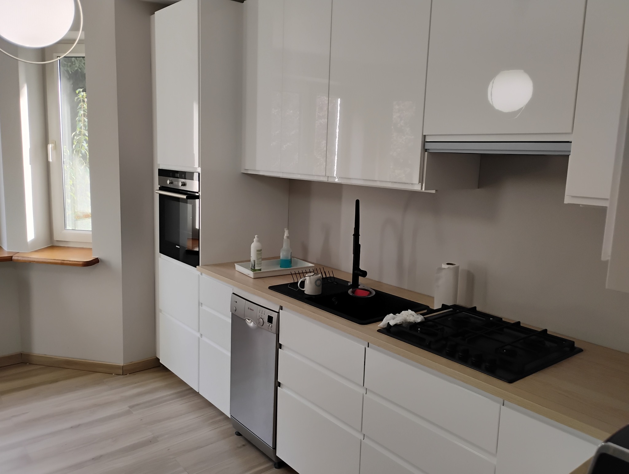 Nowoczesna kuchnia z białymi, błyszczącymi szafkami, drewnianym blatem, czarnym zlewem z wysoką baterią, płytą gazową i wbudowaną zmywarką Bosch.