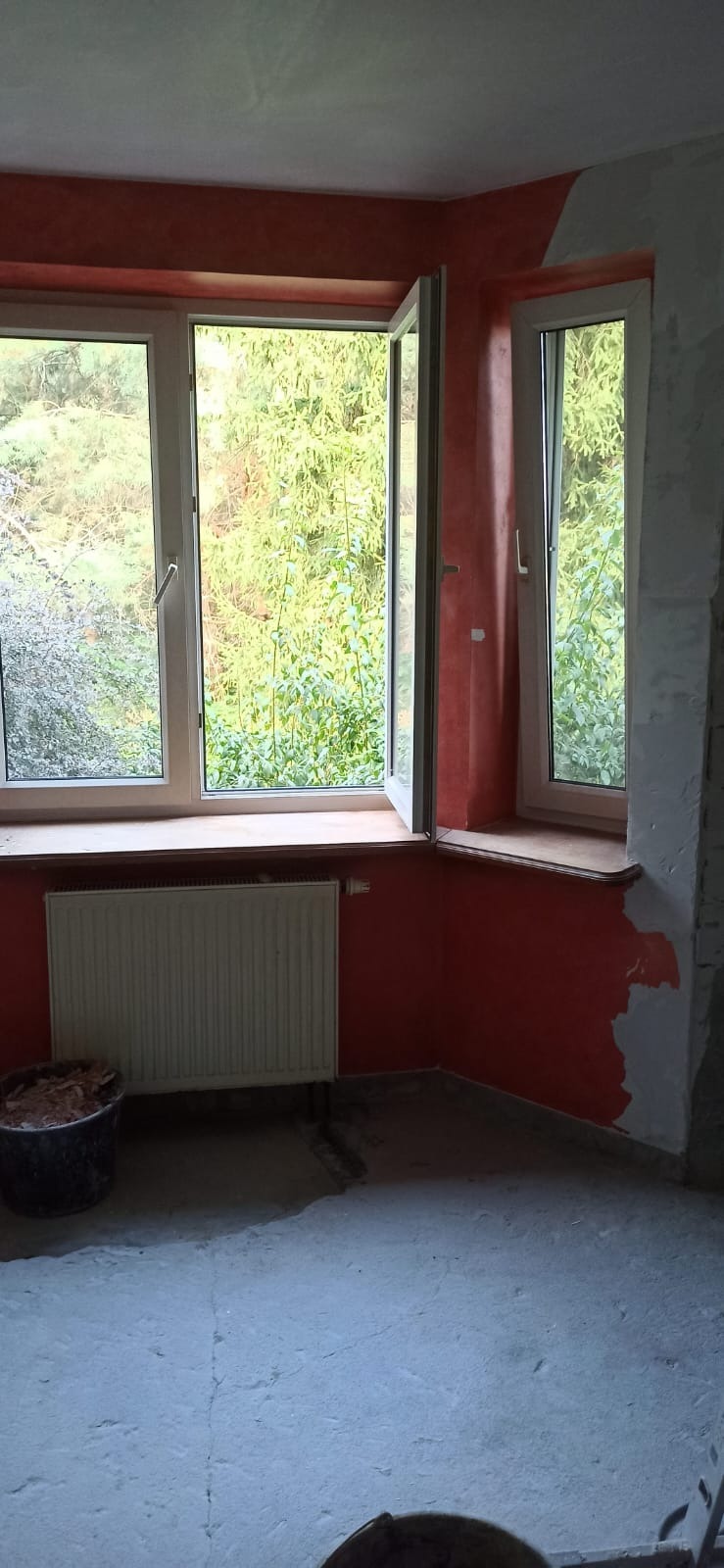 Pokój w trakcie malowania na kolor terakoty, widoczne dwa okna z zielenią na zewnątrz, grzejnik i wiadro z narzędziami malarskimi.