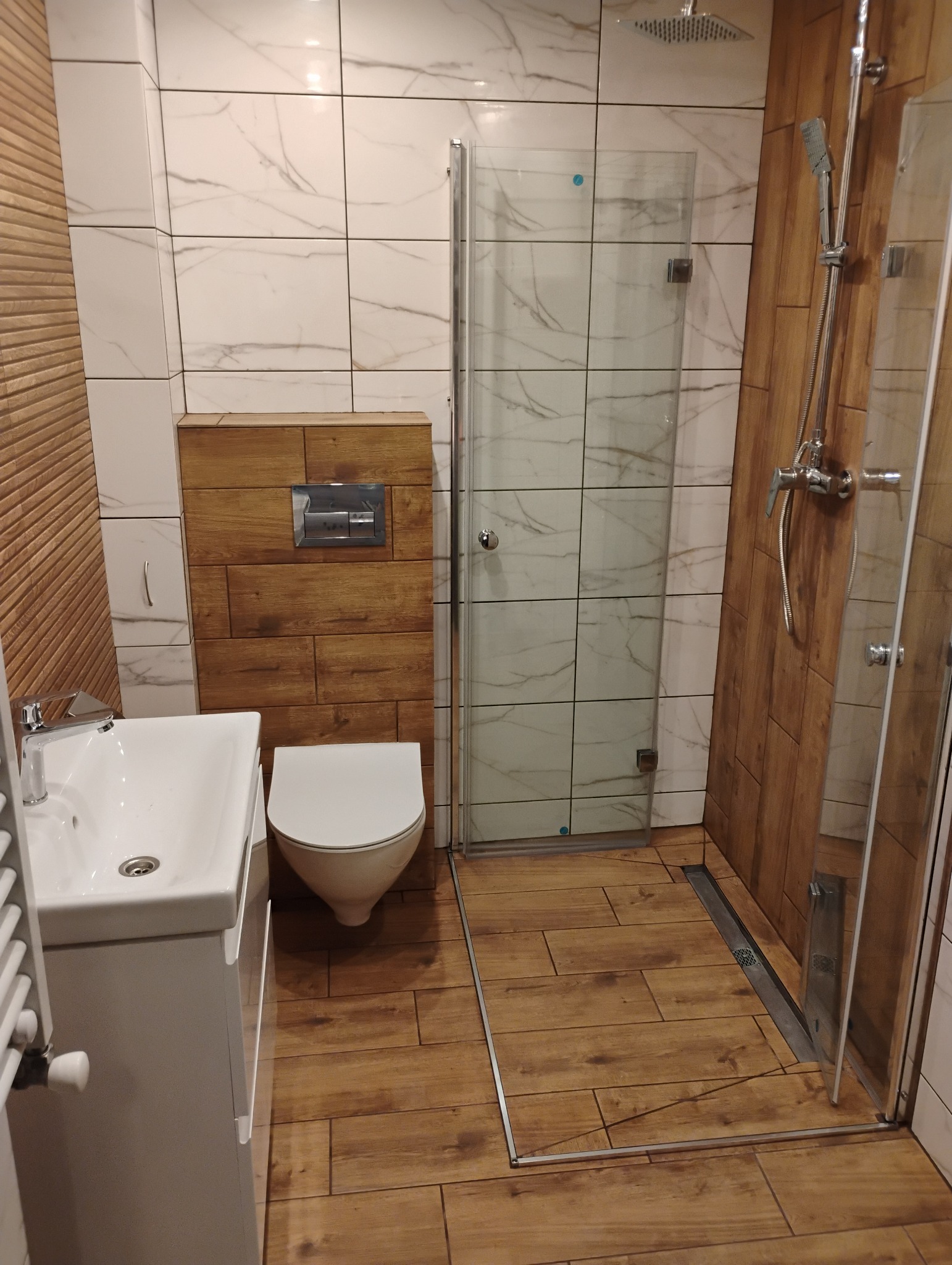 Nowoczesna łazienka z kabiną prysznicową typu walk-in, umywalką z szafką, podwieszaną toaletą w drewnianej obudowie i płytkami imitującymi marmur na ścianach.