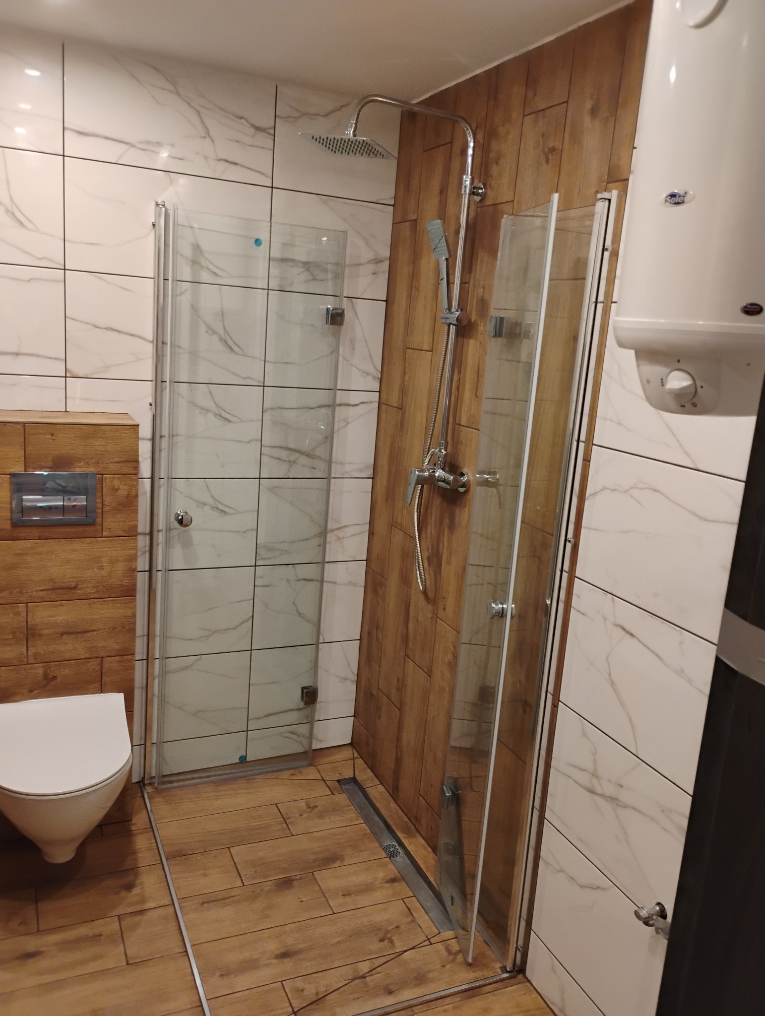 Nowoczesna łazienka po remoncie z kabiną prysznicową typu walk-in, drewnopodobnymi płytkami na podłodze i ścianach, oraz białymi płytkami imitującymi marmur na pozostałych ścianach, widoczny...