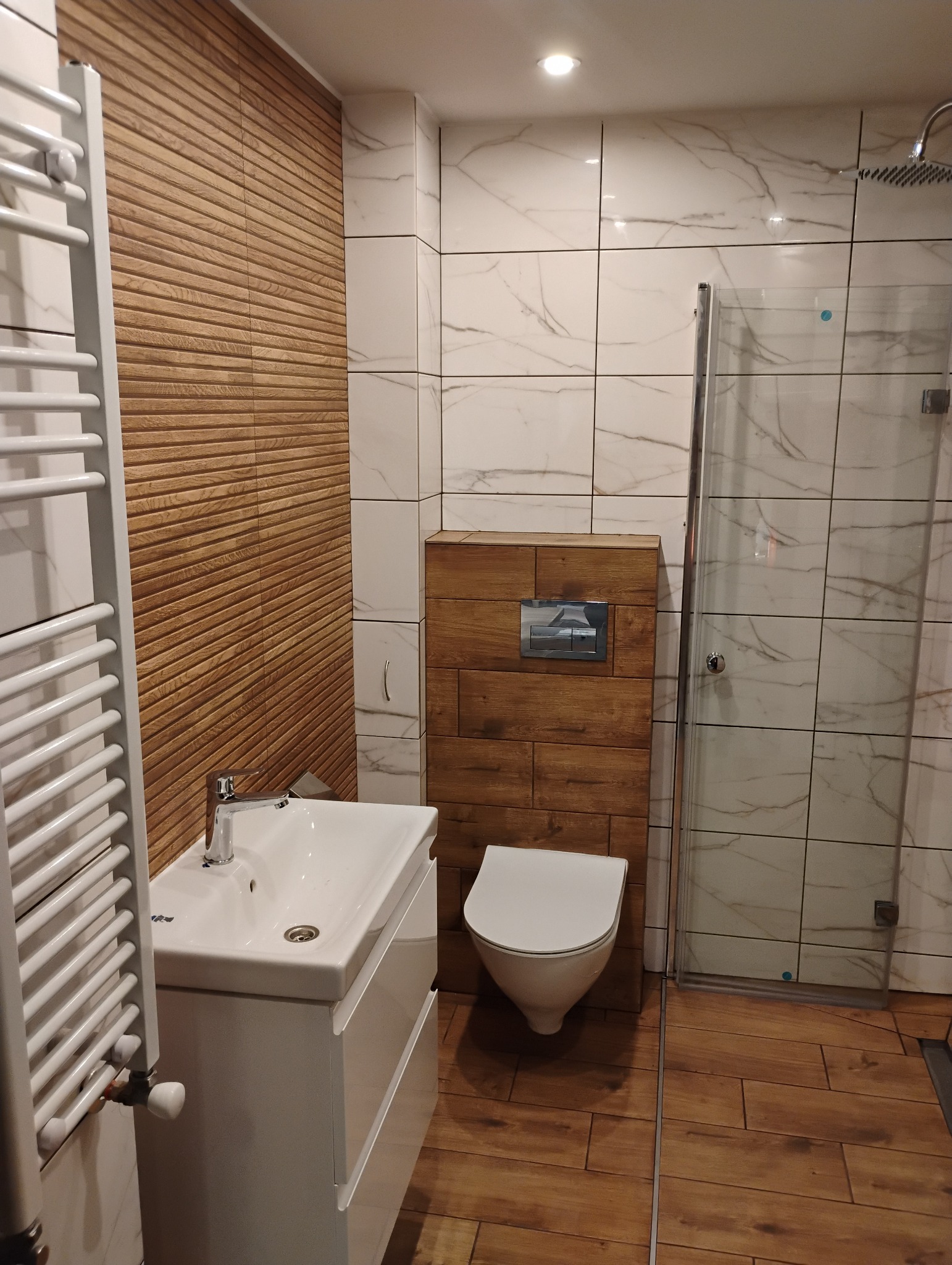 Nowoczesna łazienka z białymi marmurkowymi płytkami, drewnopodobną ścianą z poziomych paneli, wiszącą toaletą i umywalką z szafką, grzejnikiem drabinkowym oraz kabiną prysznicową.
