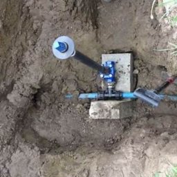 Julian usługi hydrauliczne - Niebieski zawór z czarną rurą i niebieskim przewodem, zamontowany na betonowych blokach w wykopie ziemnym.