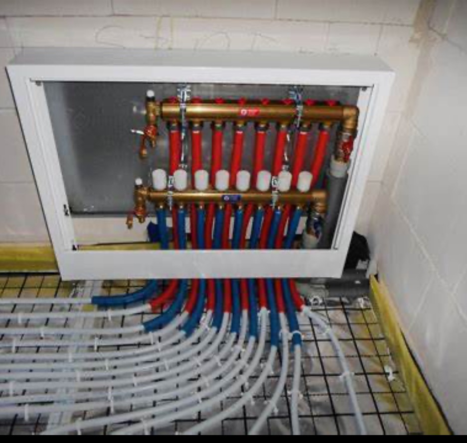 Rozdzielacz ogrzewania podłogowego w białej skrzynce, z widocznymi czerwonymi i niebieskimi rurami, wychodzącymi na siatkę montażową ogrzewania podłogowego.