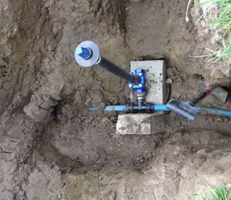 Niebieski zawór z czarną rurą i niebieskim przewodem, zamontowany na betonowych blokach w wykopie ziemnym.