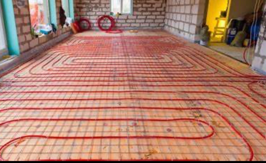 Czerwone rury ogrzewania podłogowego ułożone na siatce w surowym stanie, widoczne niedokończone ściany z cegły.