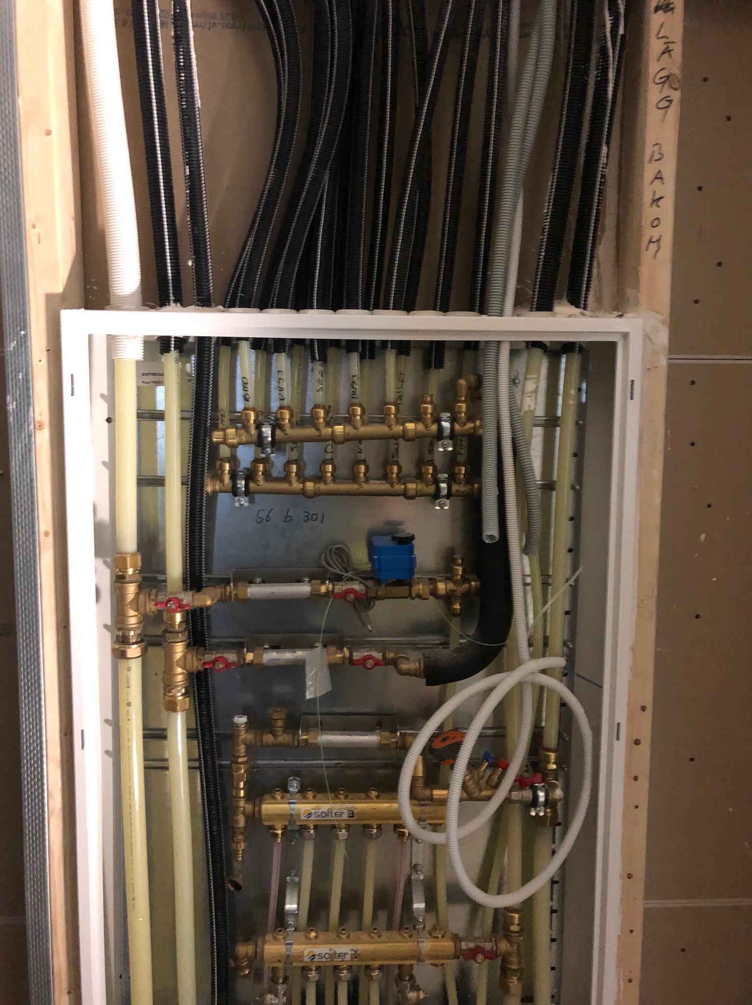 Rozdzielacz ogrzewania podłogowego w metalowej szafce, widoczne rury PEX, zawory termostatyczne z czerwonymi pokrętłami i liczne przewody doprowadzające, tło stanowi surowa ściana z widocznymi...