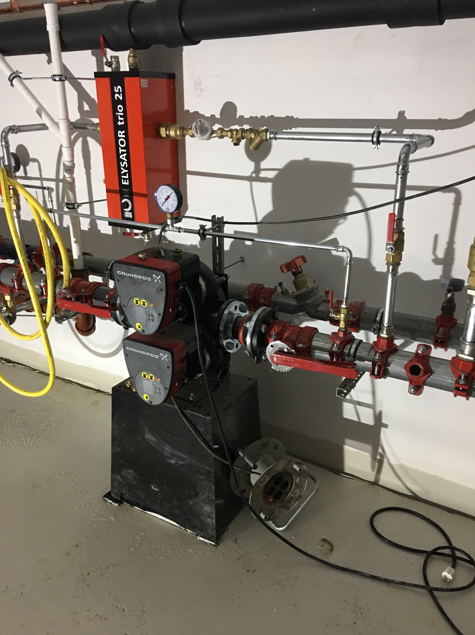 Kompleksowa instalacja grzewcza z dwoma pompami Grundfos, czerwonymi rurami i zaworami, oraz urządzeniem Elysator Trio 25, zamontowana w pomieszczeniu technicznym z betonową podłogą i widocznymi...