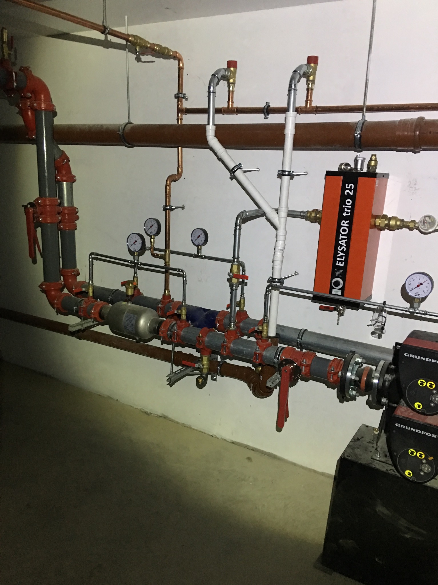 Złożony system rur grzewczych z zaworami, manometrami i pompą Grundfos w piwnicy, widoczny osad na ścianie.