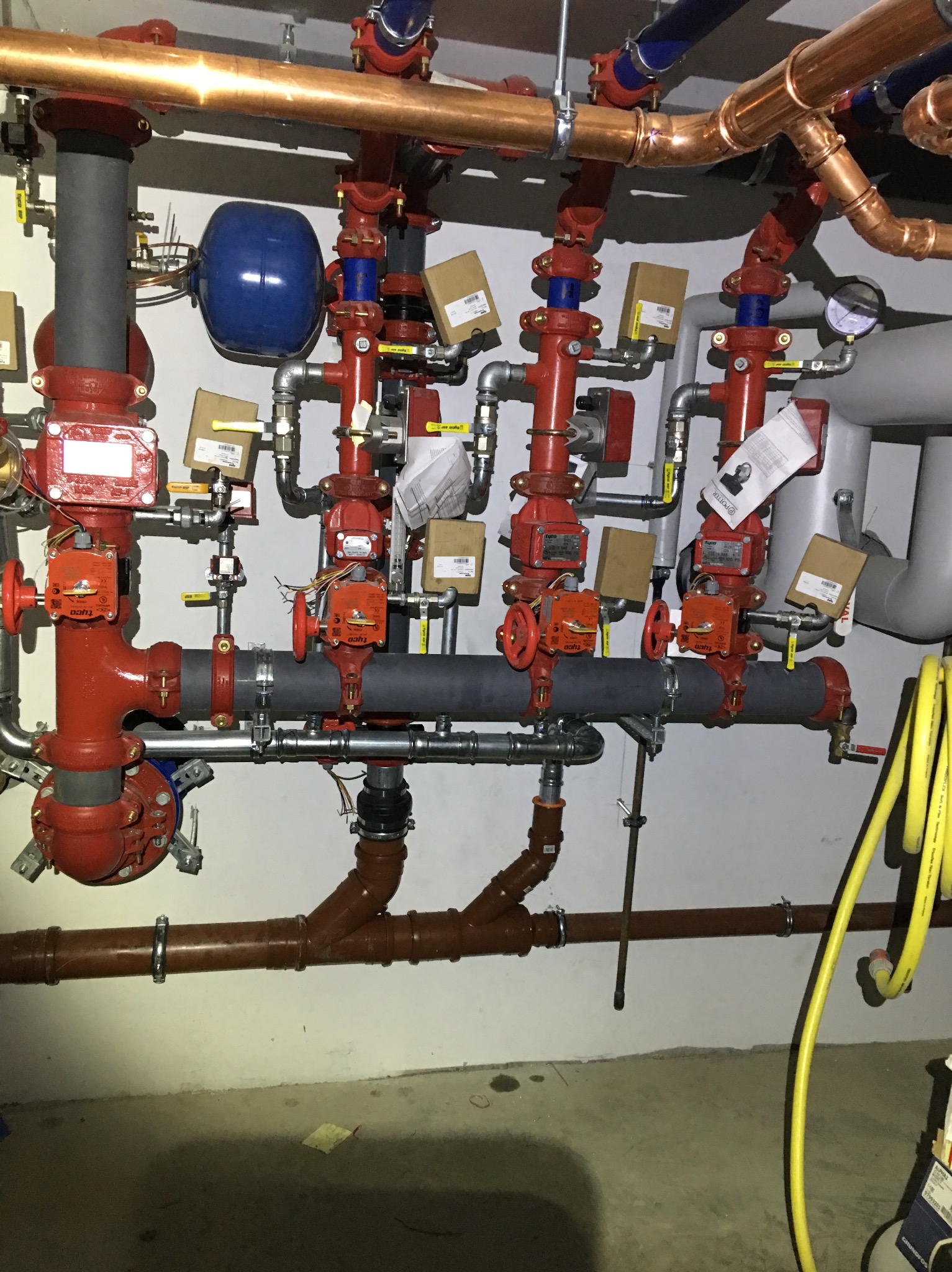 Złożona instalacja rur miedzianych i stalowych z czerwonymi zaworami, manometrem i niebieskim zbiornikiem wyrównawczym na tle białej ściany.