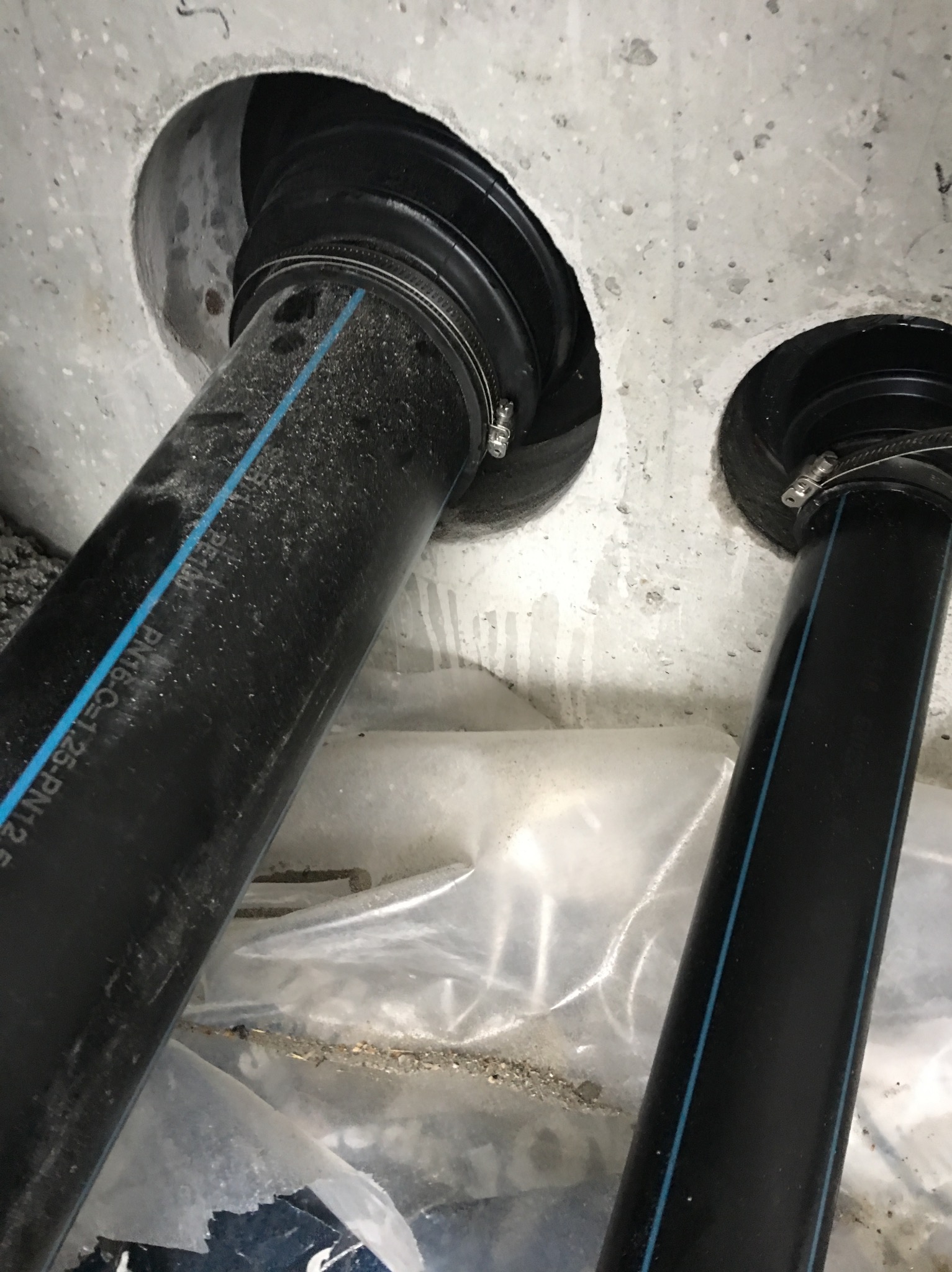 Dwie czarne rury z niebieskim paskiem wprowadzone do betonowej ściany, zabezpieczone metalowymi obejmami, widok z dołu na etapie instalacji.