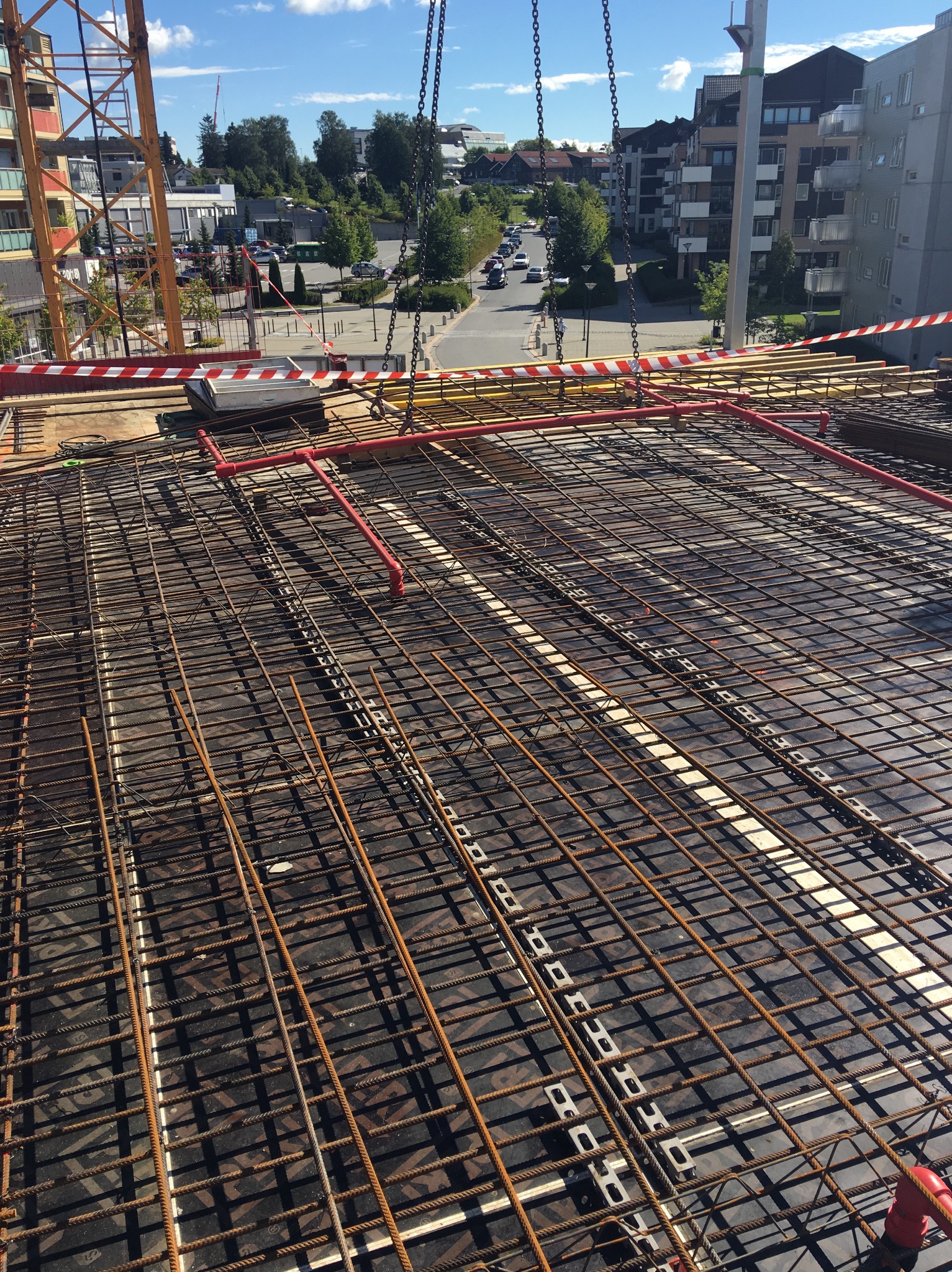 Widok z góry na siatkę zbrojeniową na budowie z rurami w kolorze czerwonym, w tle budynki mieszkalne i dźwig budowlany.