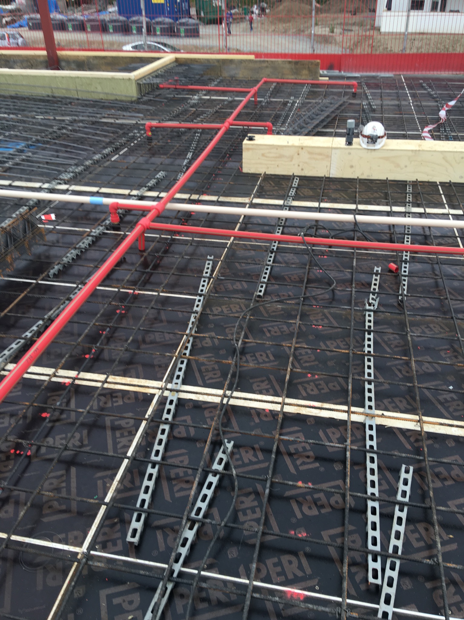 Widok z góry na siatkę zbrojeniową z zamontowanymi rurami grzewczymi w kolorze czerwonym i białym, przygotowaną do zalania betonem, na budowie. Widoczny kask ochronny i poziomica na belce drewnianej.