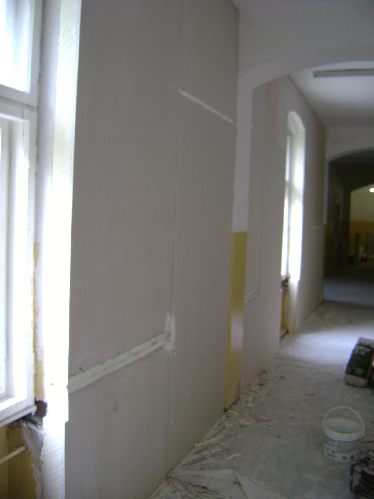 Montaż płyt izolacyjnych na ścianie w korytarzu budynku, widoczne okna i ślady kleju na podłodze.