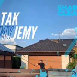 Mężczyzna w czarnej koszulce i czapce czyści dach domu z brązową dachówką za pomocą długiej lancy, w tle widoczny budynek z panelami fotowoltaicznymi, na pierwszym planie drewniany płot...