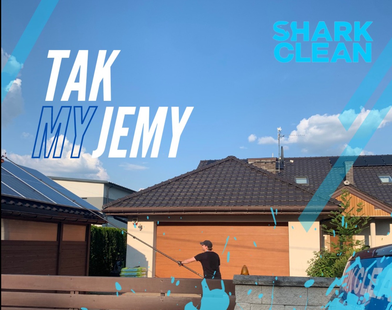 Mężczyzna w czarnej koszulce i czapce czyści dach domu z brązową dachówką za pomocą długiej lancy, w tle widoczny budynek z panelami fotowoltaicznymi, na pierwszym planie drewniany płot...