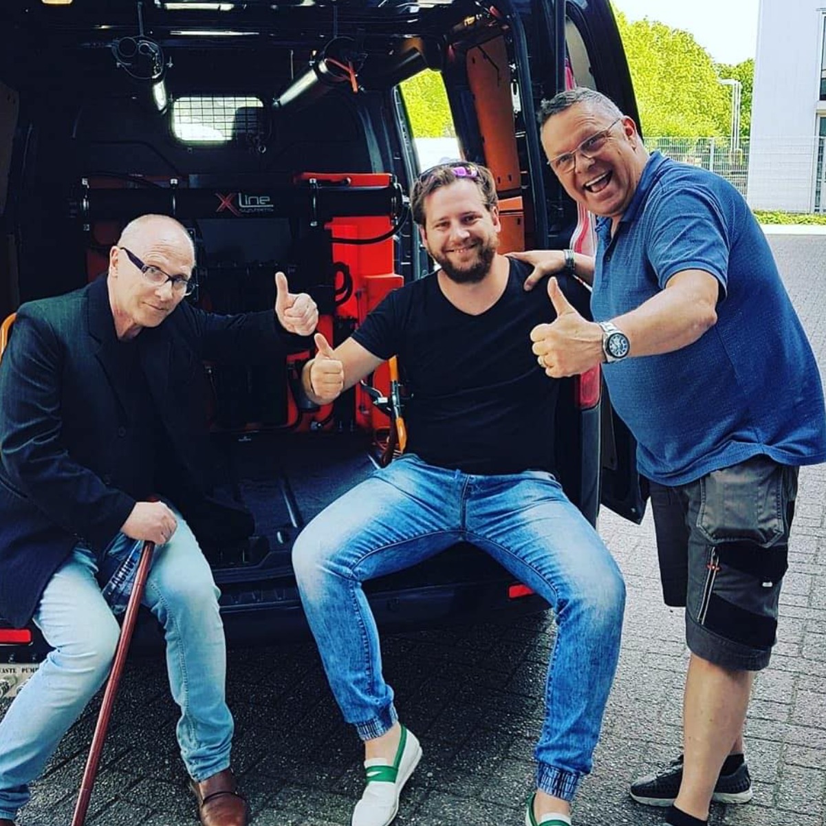 Trzech mężczyzn pozuje z podniesionymi kciukami przed otwartym bagażnikiem samochodu dostawczego z zamontowanym sprzętem do prac na wysokości.