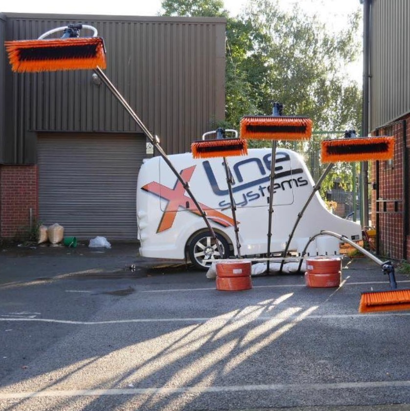 Specjalistyczny pojazd z wysięgnikami i szczotkami do czyszczenia powierzchni na zewnątrz budynku, zaparkowany na placu z widocznymi zacienieniami.