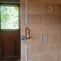 Instalacja elektryczna w surowym domu: kable w ścianie z cegły, puszki instalacyjne. Widoczne okno z zielenią na zewnątrz. Niebieska rura karbowana na dole.