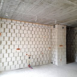 SPEC Grzegorz Piwowarski - Surowy stan: białe ściany z betonu komórkowego, widoczne przewody elektryczne i puszki instalacyjne, sufit betonowy. W tle widoczne wejście do pomieszczenia.