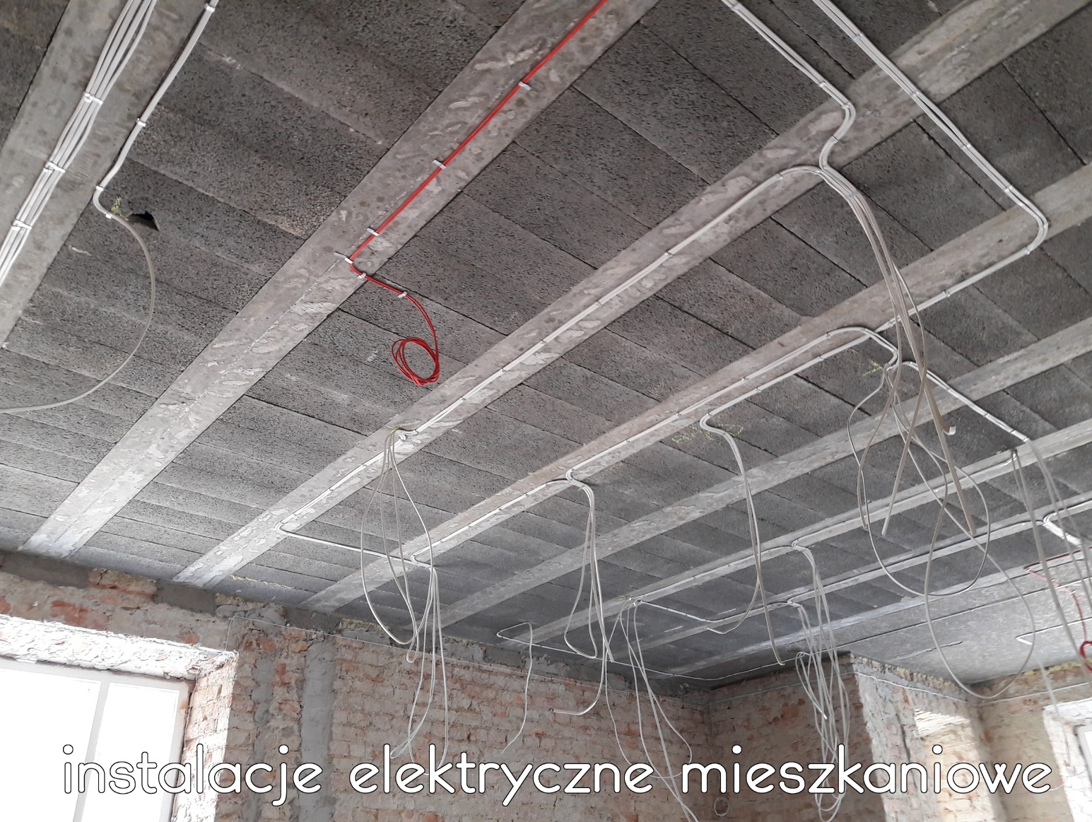 Surowy strop z szarego betonu z widocznymi przewodami elektrycznymi w białych rurkach i jednym czerwonym, przymocowanymi do belek stropowych, zwisające przewody w nieukończonym wnętrzu.
